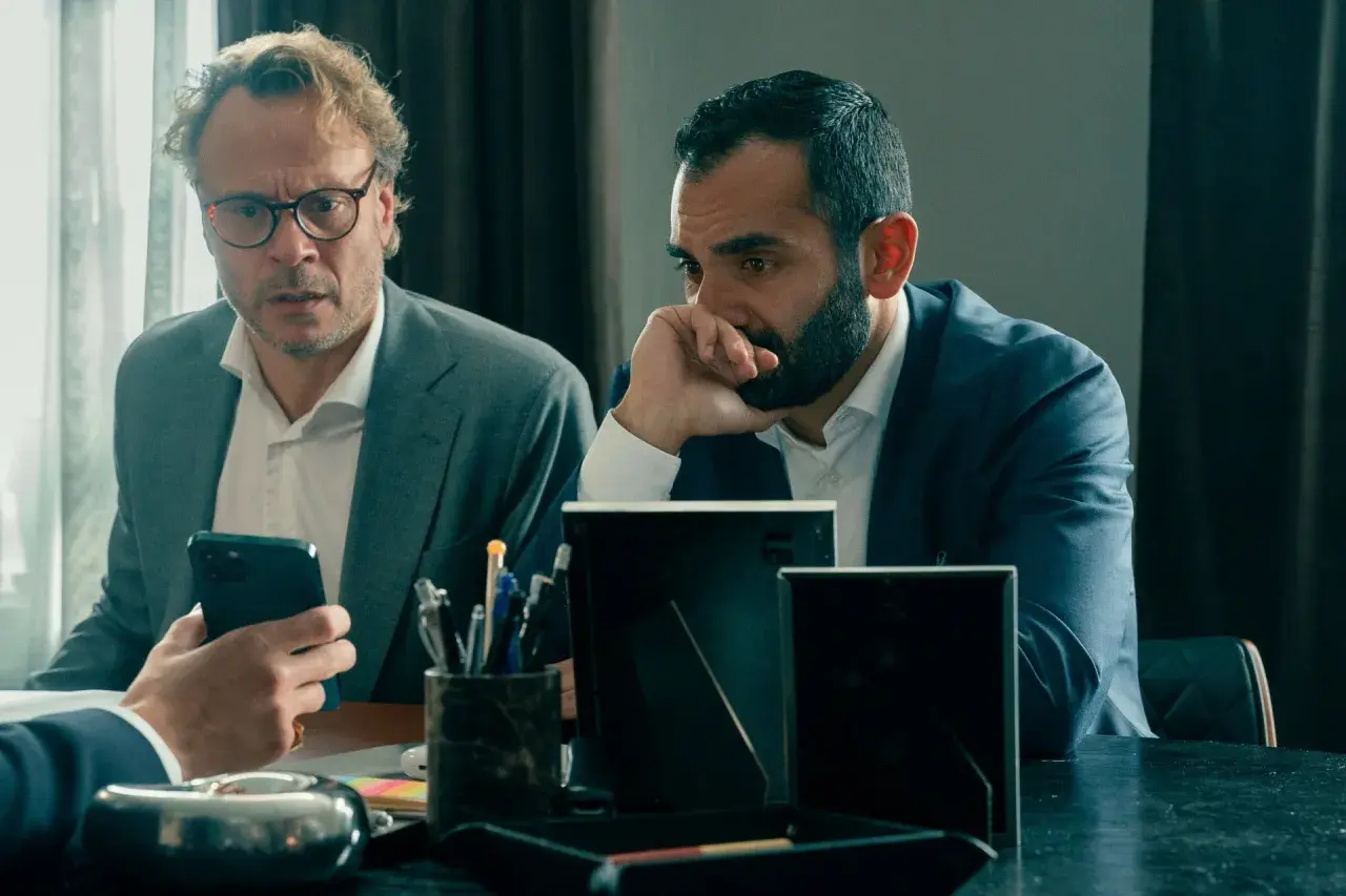 Två män i kostym sitter vid ett bord. Den ena mannen, med glasögon, tittar på sin mobiltelefon. Den andra mannen, med skägg, funderar djupt. Scen från "hundarna" på SVT.