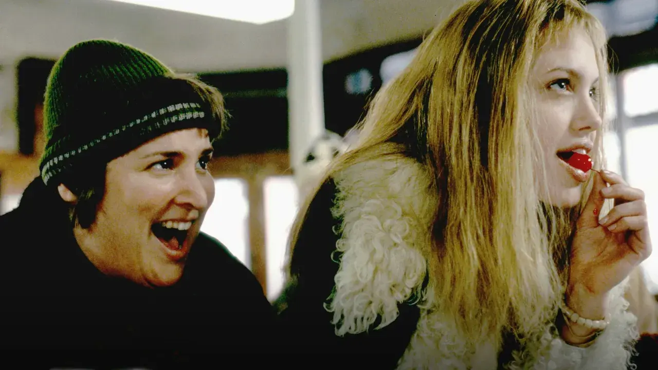 En glad kvinna med grön mössa och en annan kvinna med blont hår som äter ett körsbär. Scen från "Girl, Interrupted" (Stulna år).