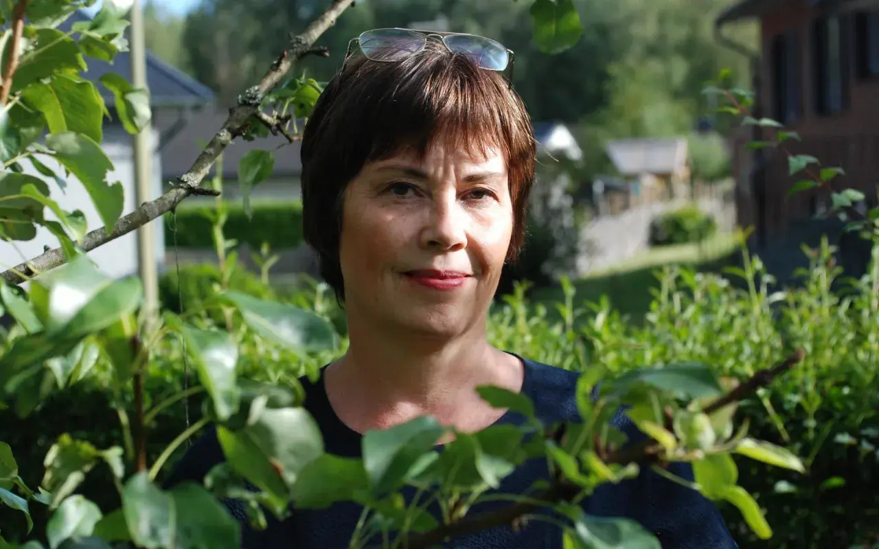 Anna Bratt, en kvinna med glasögon på huvudet, ler mot kameran i en lummig trädgård.