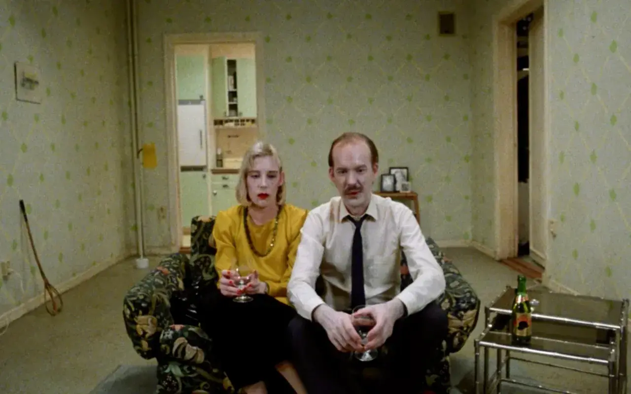 Ett par sitter i en soffa, en kvinna med gul tröja och en man i skjorta och slips. Kanske en scen från en Roy Andersson reklamfilm.