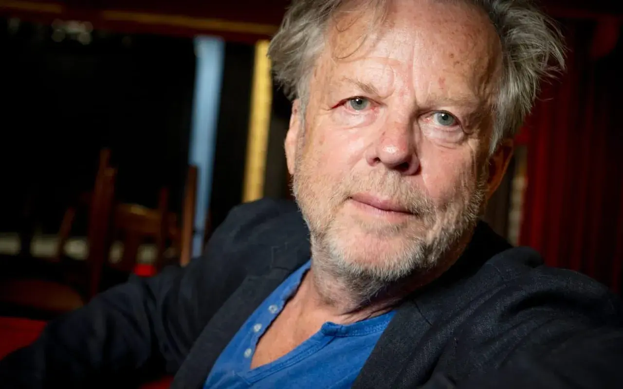 En äldre man med grått hår och skägg, klädd i kavaj och blå tröja. Han påminner om en ung Wallander.