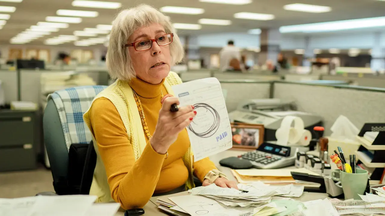 Jamie Lee Curtis som en kontorist i filmer, med glasögon och en gul polotröja, håller upp ett papper med texten "PAID".