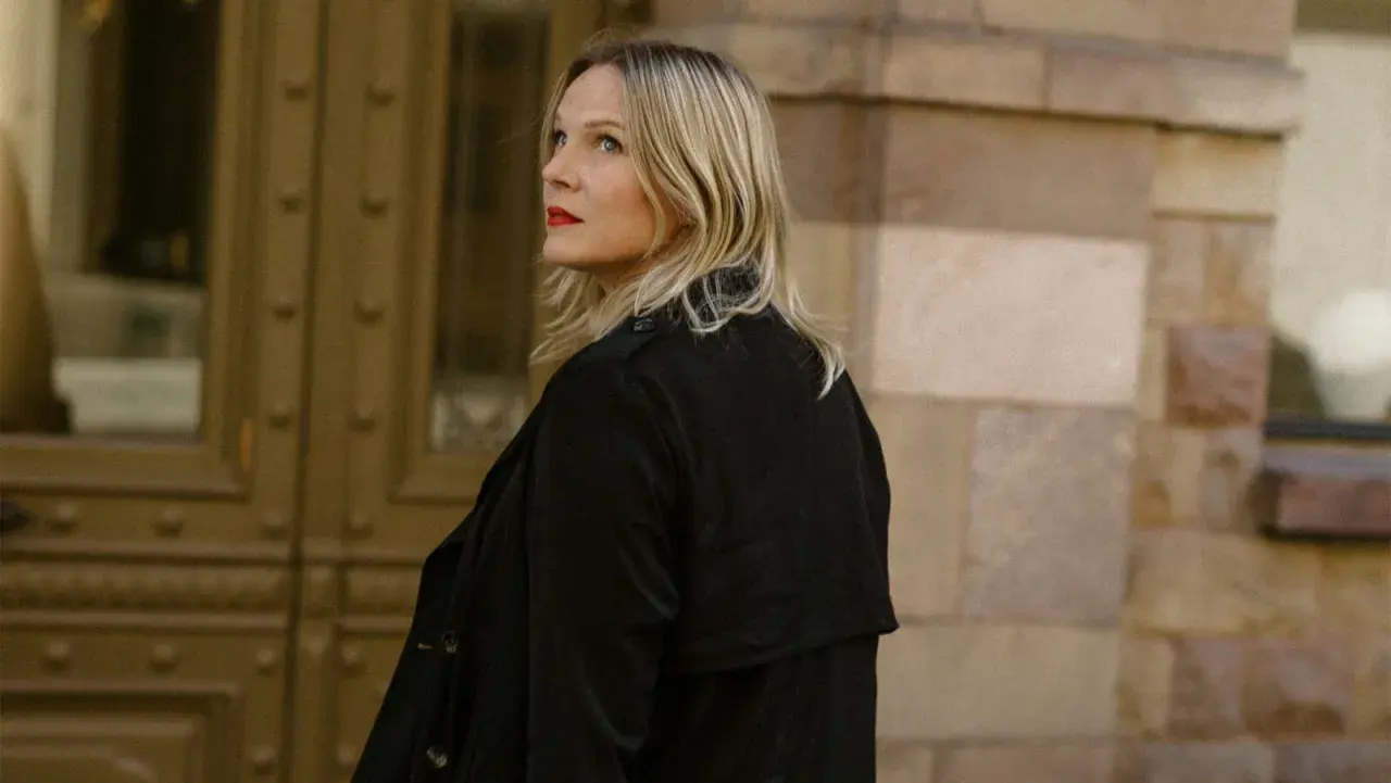 Jenny Aschenbrenner, med rött läppstift och blont hår, tittar uppåt mot en byggnad i sten.