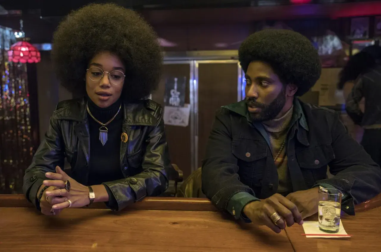 En kvinna med stor afro och en man med skägg sitter vid en bardisk. De är medlemmar i rollistan i Blackkklansman.