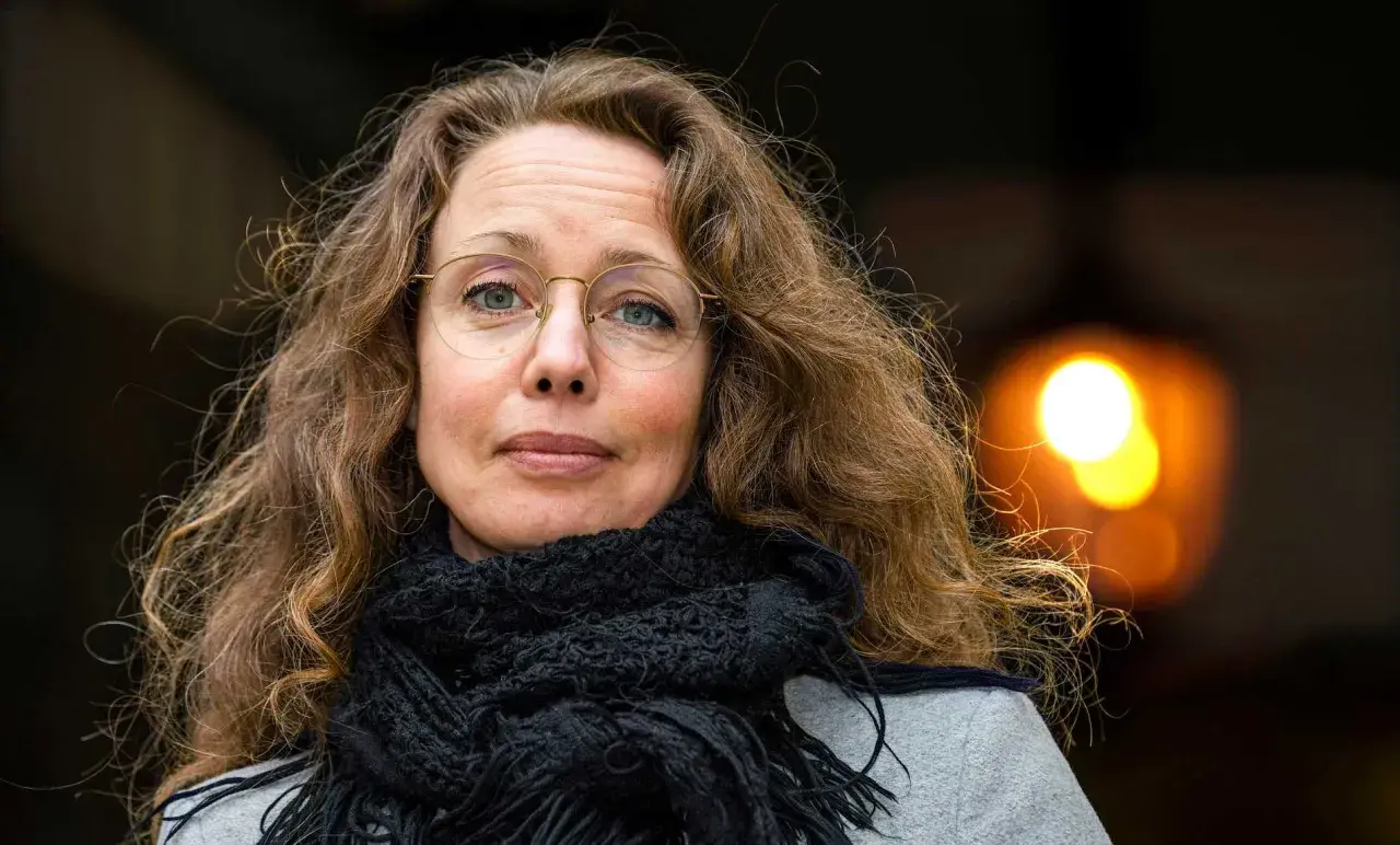 Sara Gordan dotter med sitt långa, lockiga hår och en svart halsduk. Hon bär glasögon och tittar rakt in i kameran.