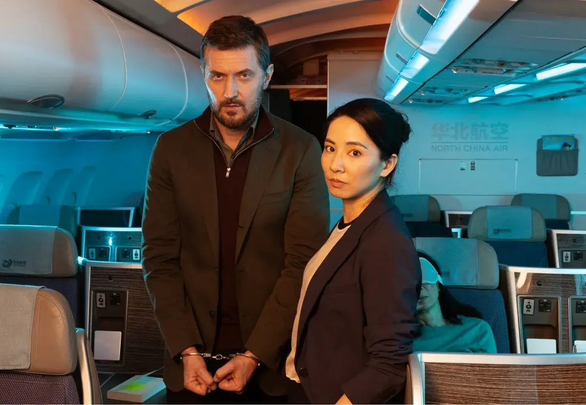 Man med handbojor och kvinna står i ett flygplan. En sovande passagerare syns i bakgrunden. Scen från konspirationen serie.