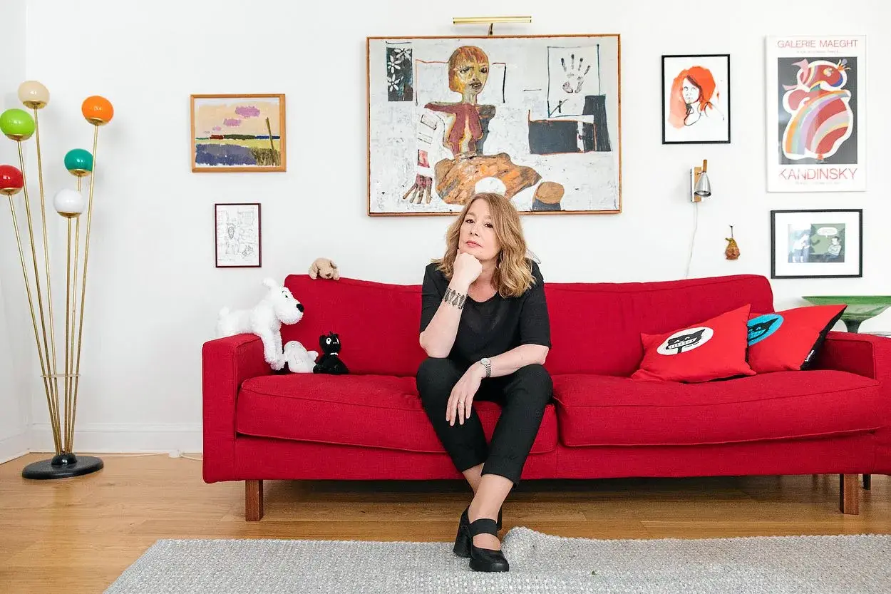 Åsa Linderborg sitter på en röd soffa, omgiven av konst och leksaker som påminner om barn.