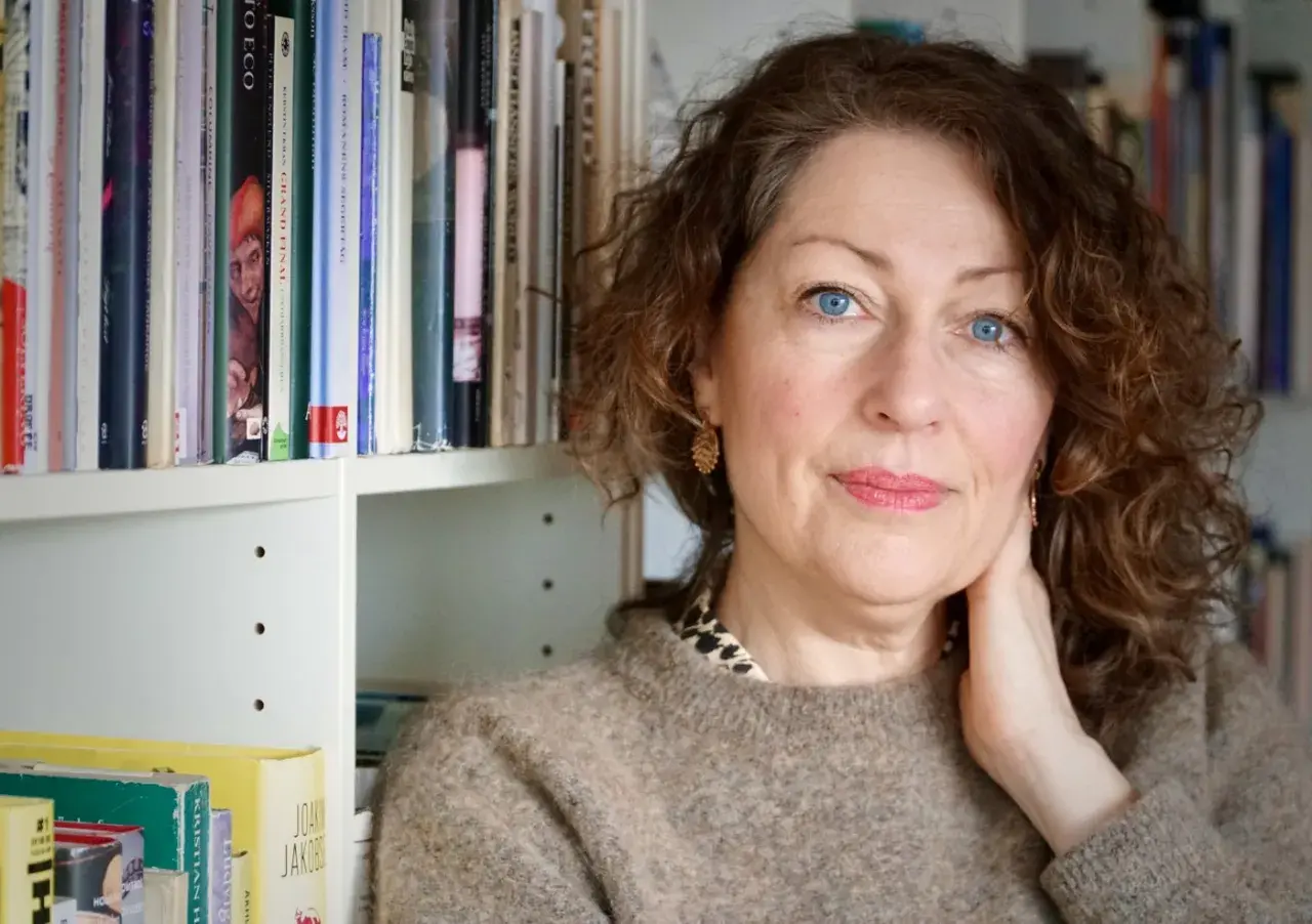 Elisabeth Åsbrink, med blå ögon och lockigt hår, sitter framför en bokhylla. Hon verkar lite sjuk, men hennes blick är klar.