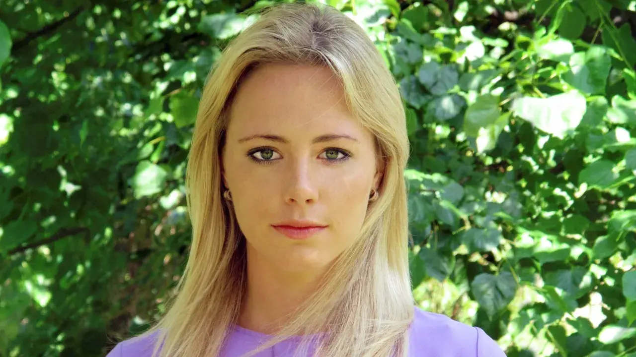Close-up portrait of a blonde woman with green eyes and a purple top, against a blurred green foliage background.