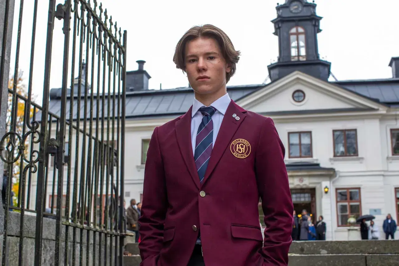En ung man i skoluniform, som påminner om unga kungligheter, står framför en ståtlig byggnad.