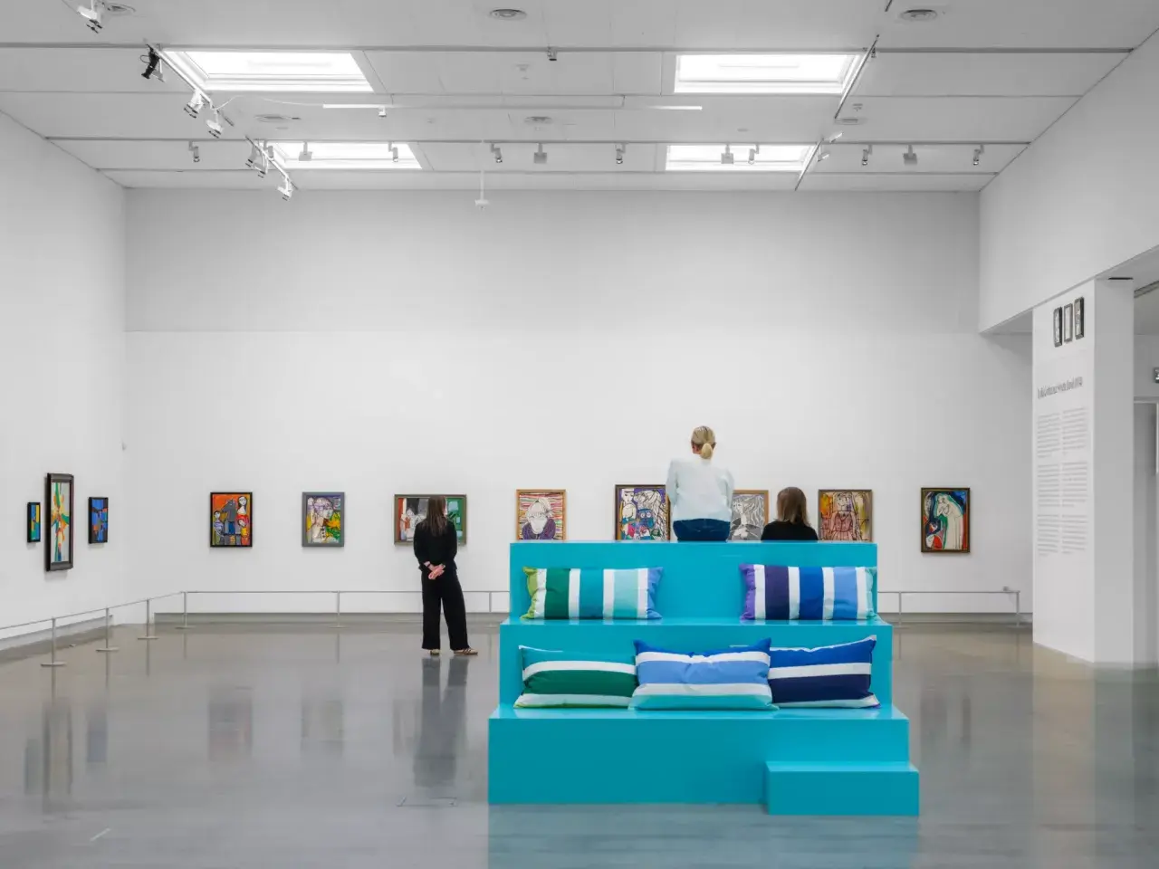 I en konsthall sitter besökare på en turkos scen med kuddar, omgivna av färgglada tavlor. Kanske är de där för att inspireras, för att vara en musa för konstnären.