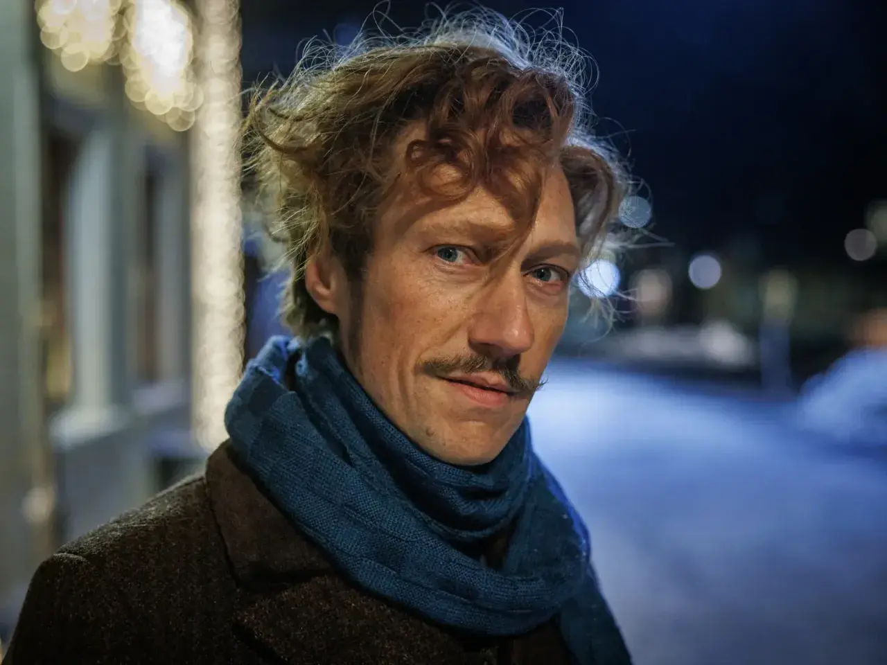 Man med rufsigt hår och mustasch, klädd i brun rock och blå halsduk. Kanske en del av rollistan i jakten på en hemlighet i vintermörkret.