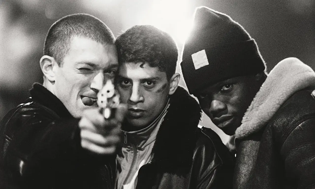 Tre unga män, en med en pistol riktad mot kameran, i en dramatisk scen från "La Haine" (Medan vi faller).