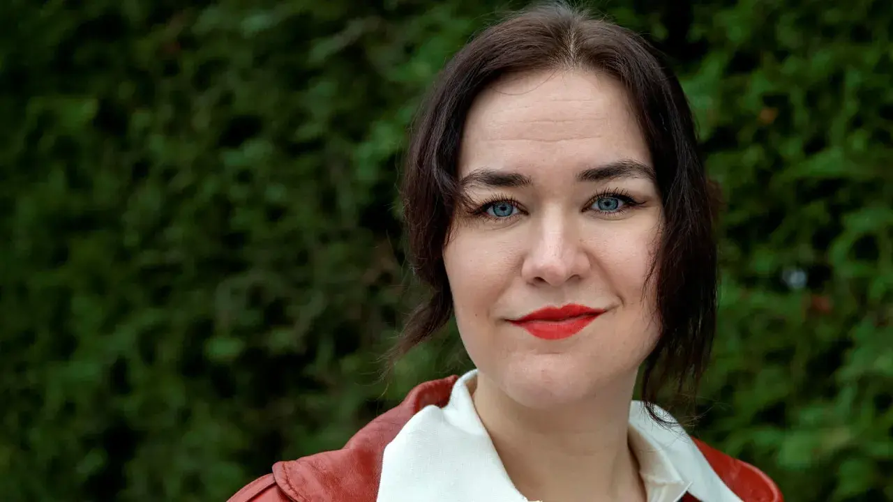 Camilla Sten med blå ögon och rött läppstift, klädd i en röd jacka, ler mot kameran.