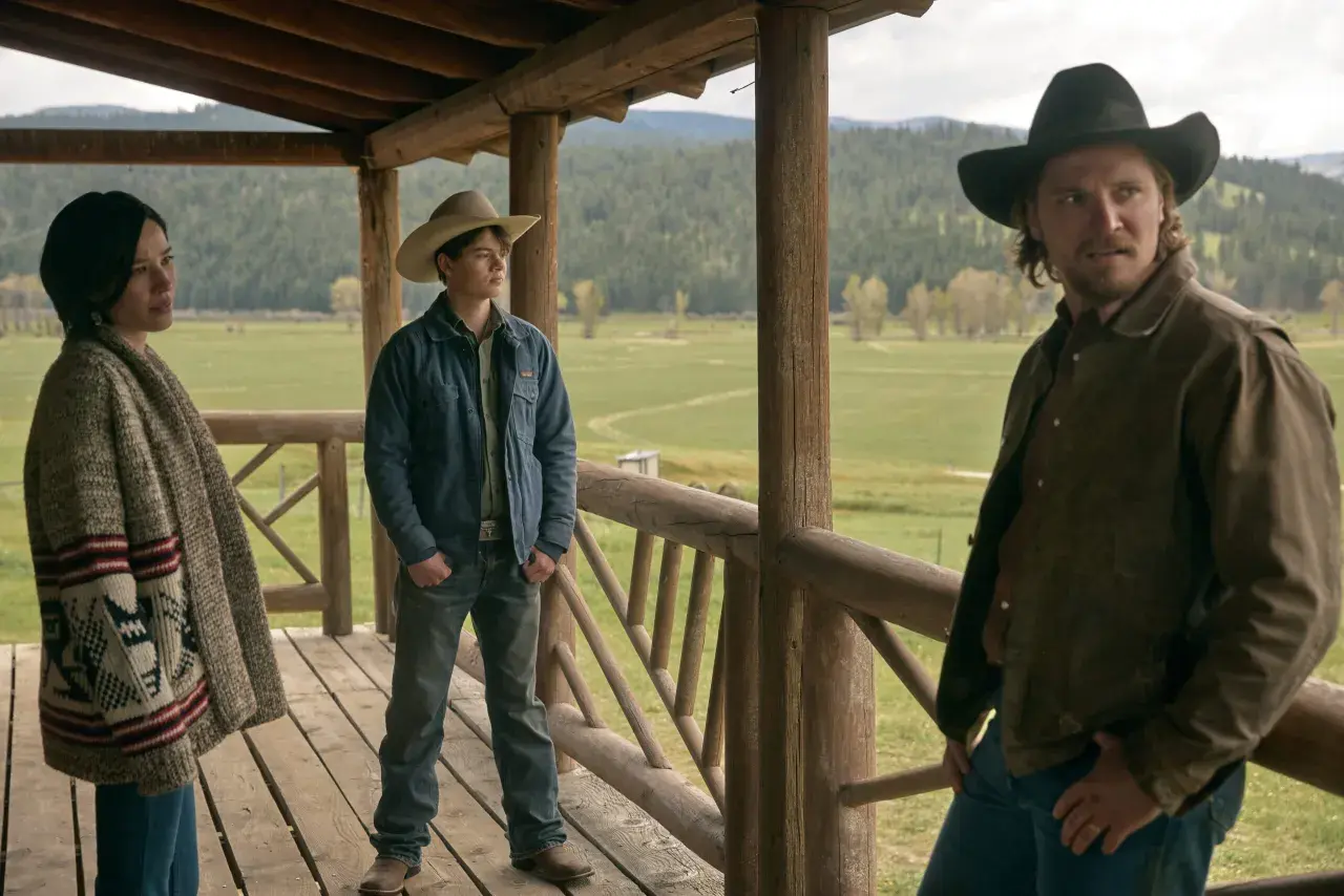 Tre personer på en veranda med berg i bakgrunden. En kvinna med mönstrad kofta, en ung man med cowboyhatt och en man med hatt och brun jacka. Scen från Yellowstone-serien.