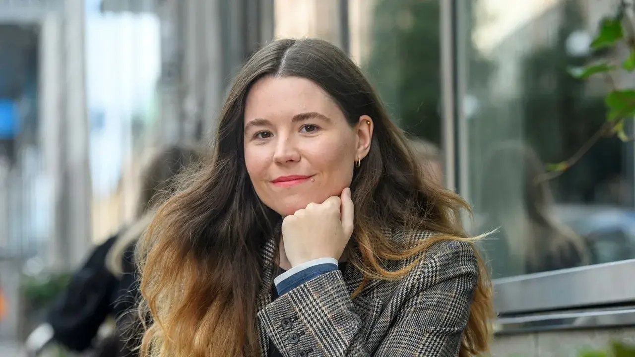 Johanna Hedman, med sitt långa, vågiga hår och en rutig kavaj, ler mot betraktaren.