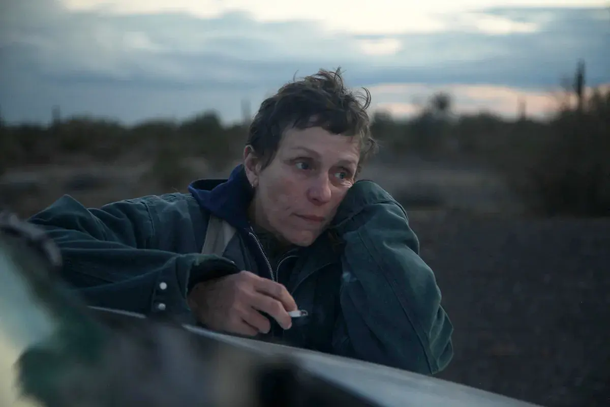 En kvinna med en cigarett i handen lutar sig mot en bil i ett öde landskap. Hon är en del av rollistan i Nomadland.