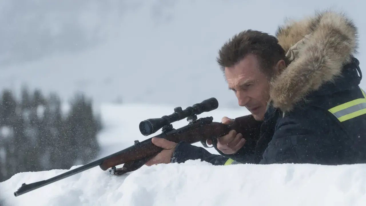 Liam Neeson i en vinterjacka med luva siktar med ett gevär i snön.