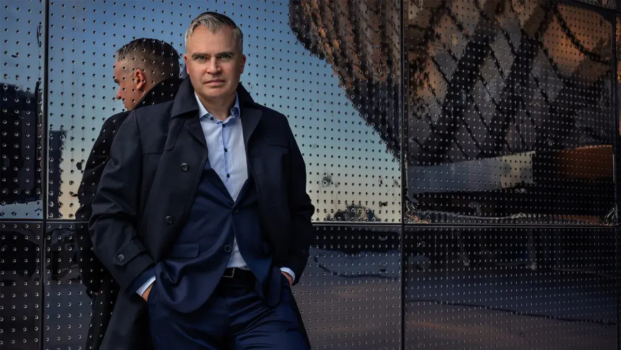 Jerker Virdborg står framför en reflekterande fasad, klädd i kostym och rock.