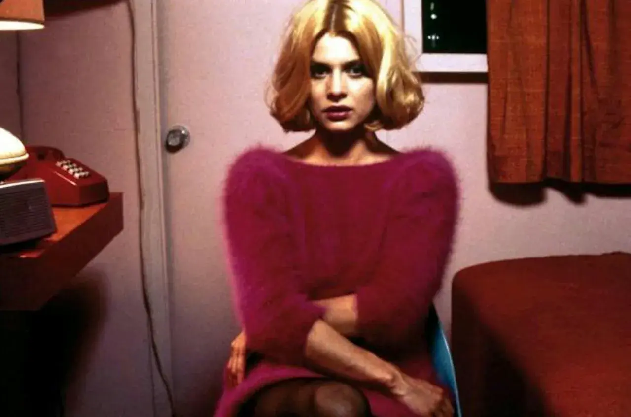 En kvinna med blont hår sitter i ett rum, kanske på ett motell. Hon bär en rosa tröja. En röd telefon syns på en hylla. Scen från "Paris, Texas".
