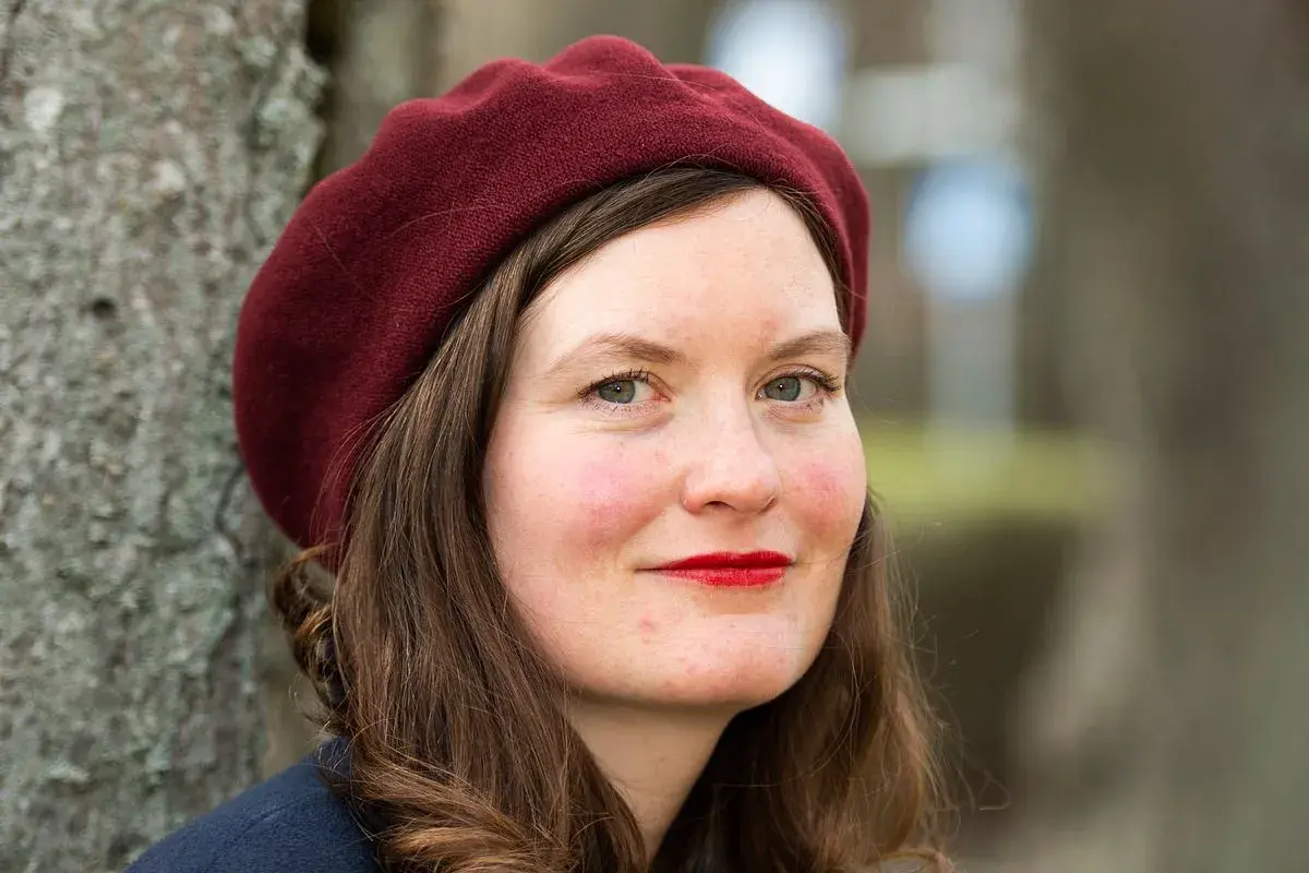 Alva Dahl, med en vinröd basker och röda läppar, ler mot kameran. Hon står utomhus vid ett träd.