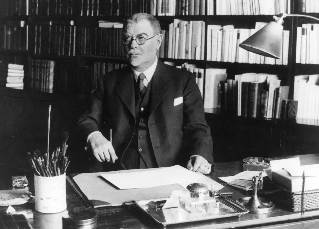 Albert Engström, med glasögon och kostym, sitter vid sitt skrivbord med böcker i bakgrunden. Penslar och bläckhorn syns på bordet.