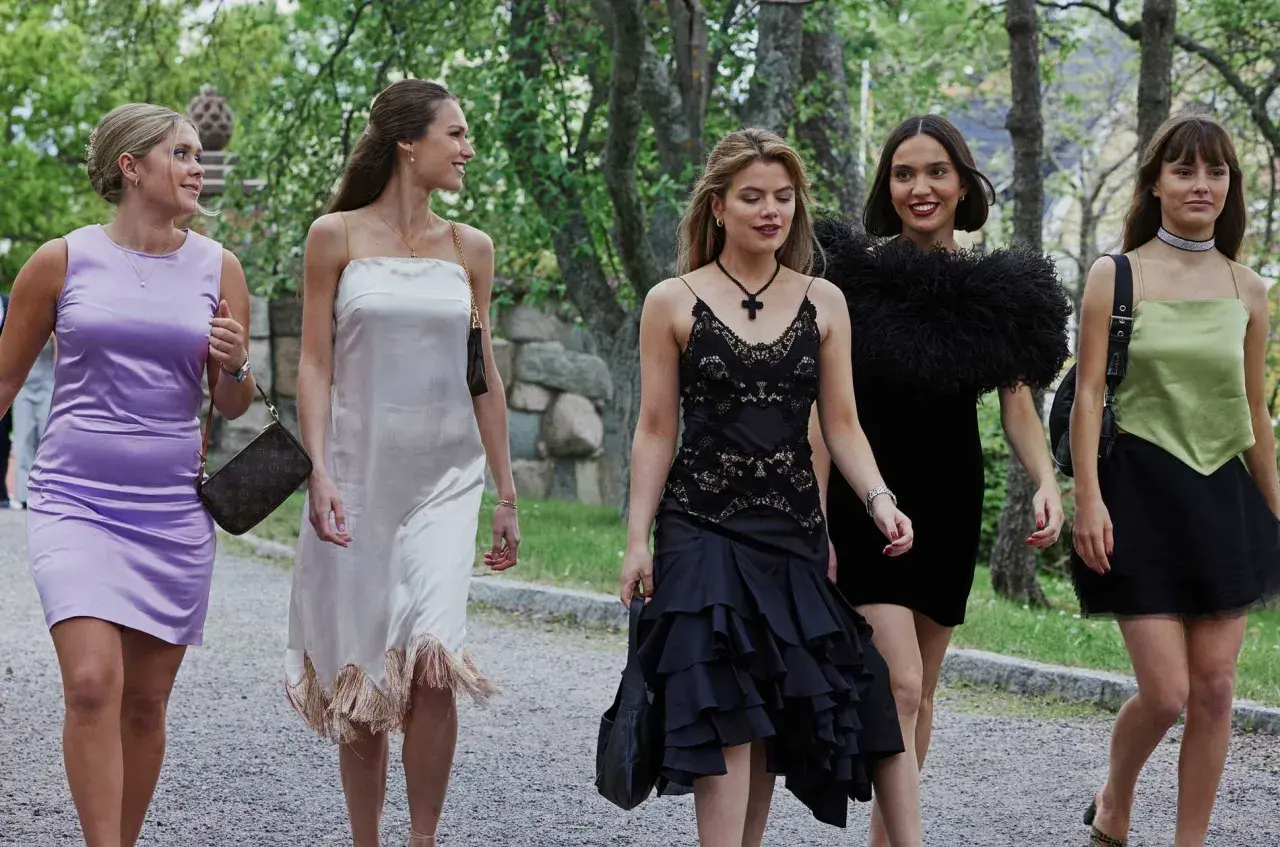 Fyra unga kvinnor i eleganta klänningar, som barracuda queens, promenerar utomhus.