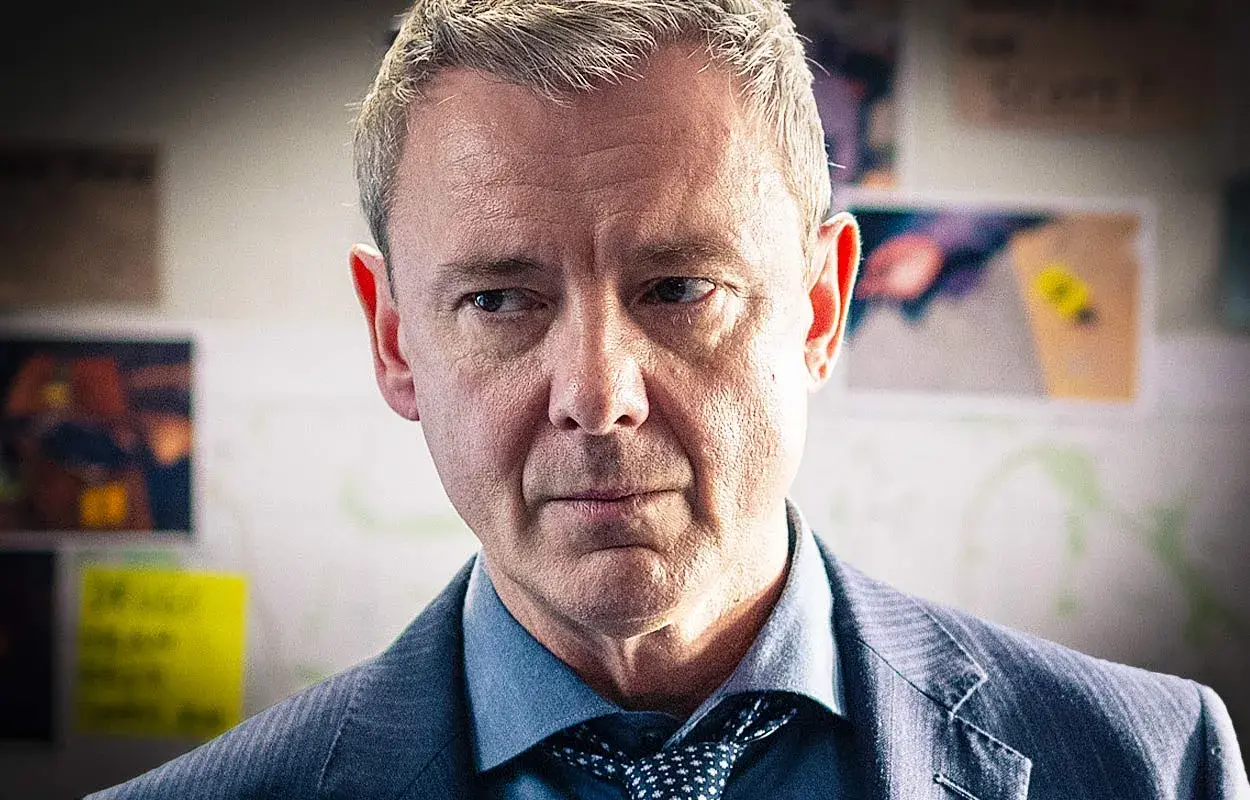 En man i kostym, med grånande hår, blickar fundersamt framåt. Han är en del av grace serie.