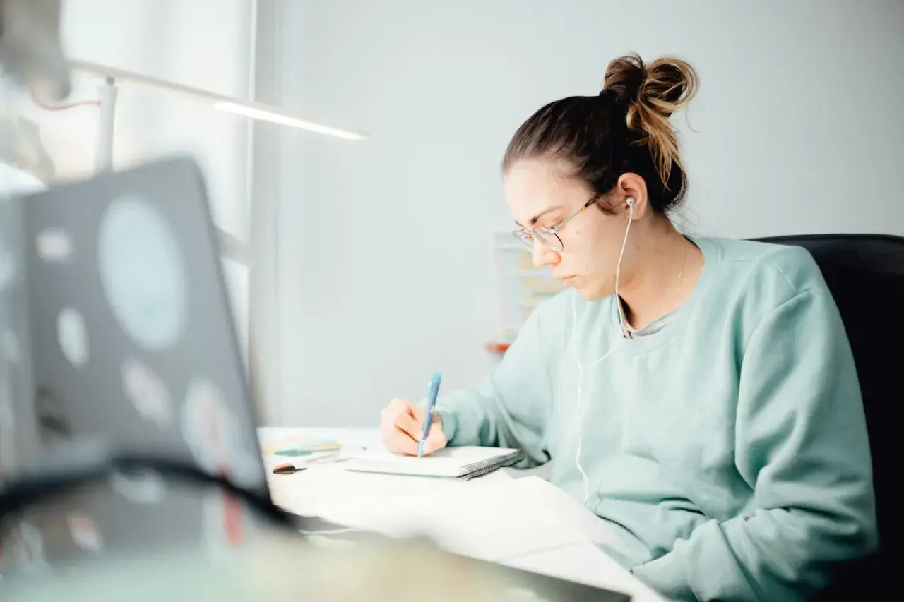 Student med hörlurar skriver i anteckningsbok vid skrivbordet, inspirerad av sitt universitetsutbildning i kreativt skrivande.