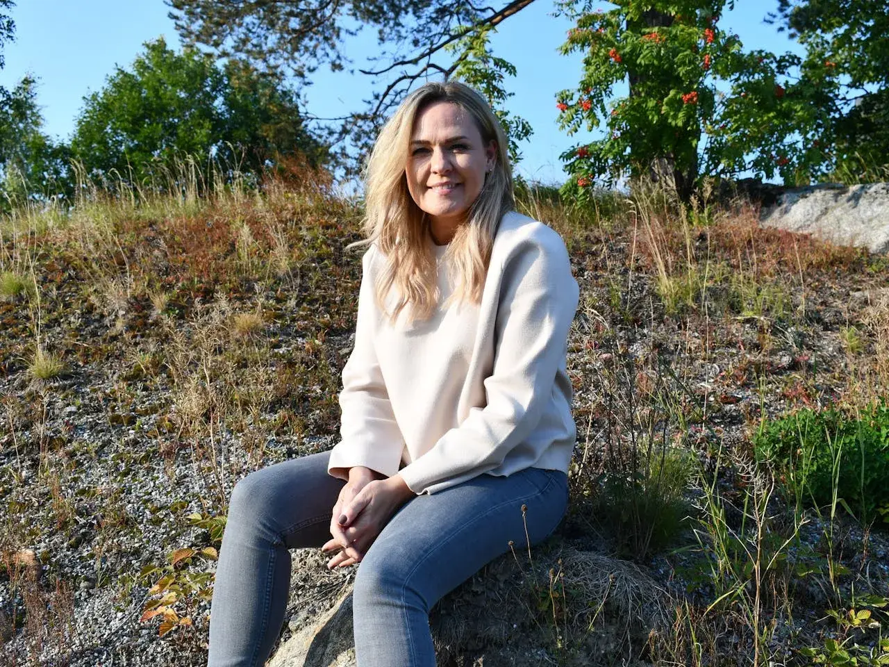 Ulrika Rolfsdotter sitter på en sten utomhus, omgiven av gräs och träd.