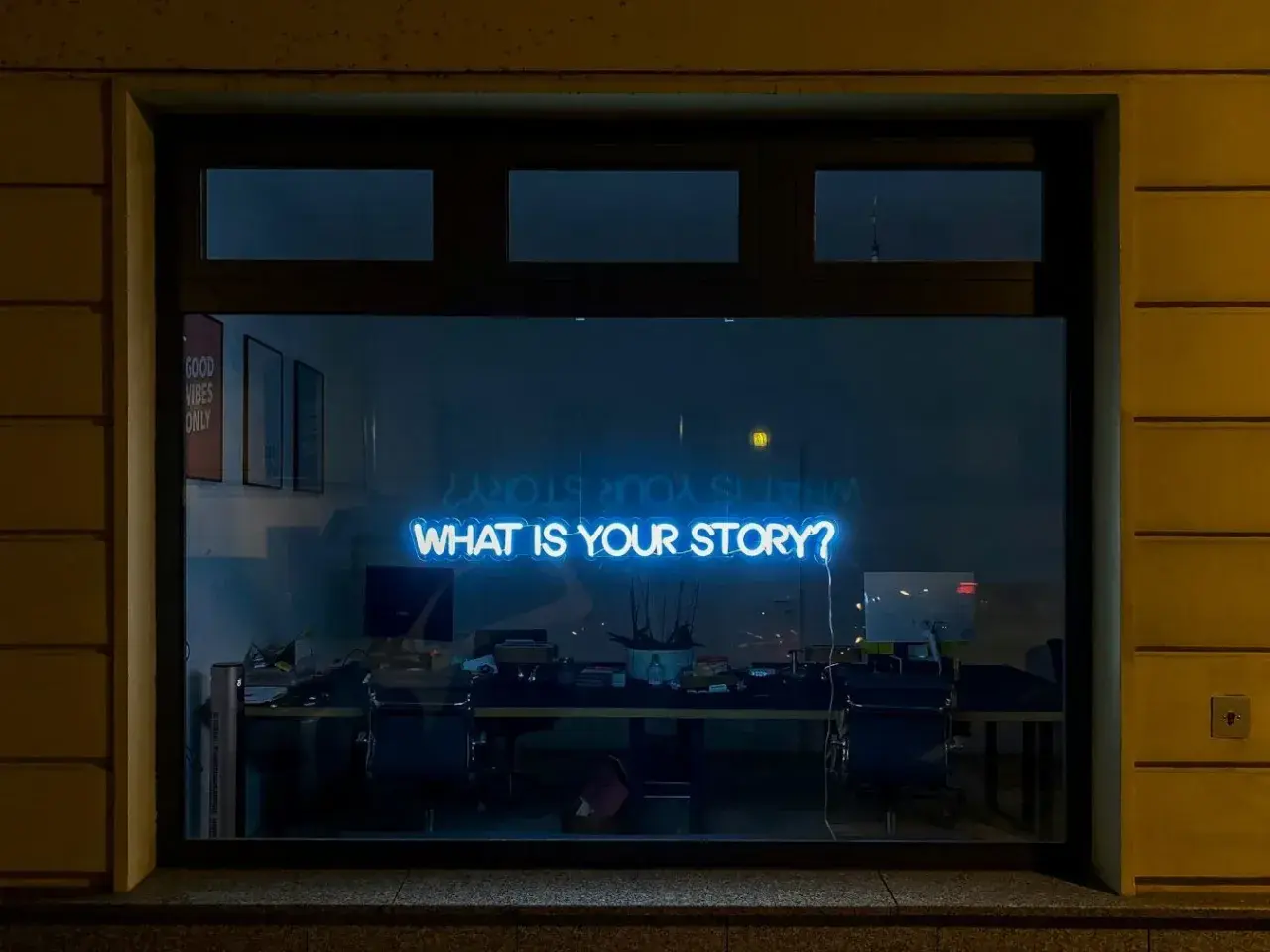 Neonskylt med texten "WHAT IS YOUR STORY?" lyser blått i ett fönster, som en inbjudan till en kurs i skrivande.