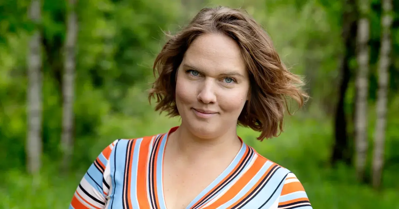 Underbara Claras leende möter kameran i en randig klänning, med en lummig skog i bakgrunden.
