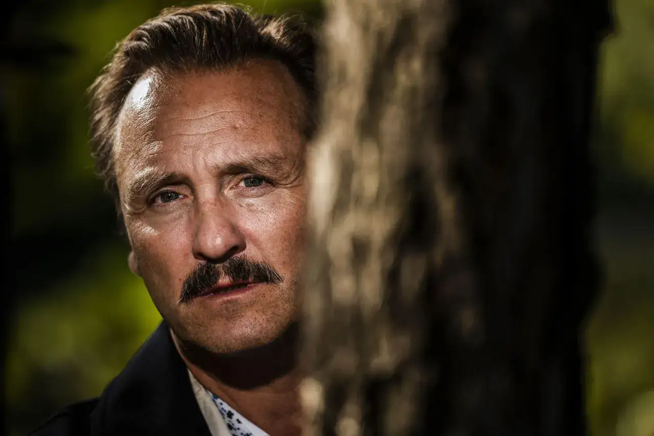 Simon Sears, med mustasch och blick som anar skogen, står bakom ett träd.