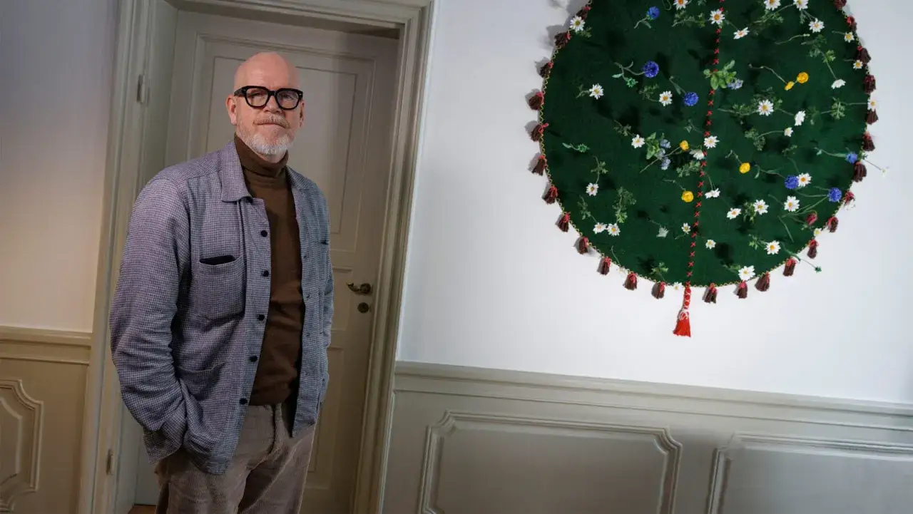 Jonas Axelsson, en man med glas&ouml;gon och sk&auml;gg, st&aring;r bredvid ett runt, gr&ouml;nt konstverk med blommor och tofsar.