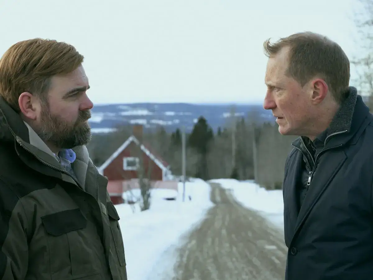 Två män står vända mot varandra i ett snöigt landskap. En väg leder upp mot ett rött hus. The shadow line mellan dem antyder en spänd situation.