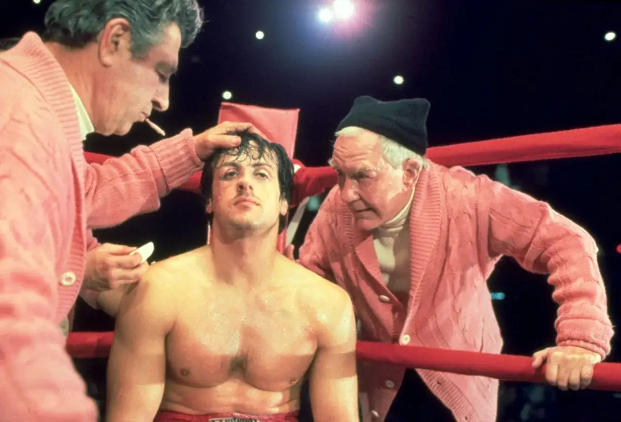 Rocky Balboa, med smuts och sår i ansiktet, får omsorg av Mickey och Paulie i ringhörnan. Rollistan i Rocky är ikonisk.
