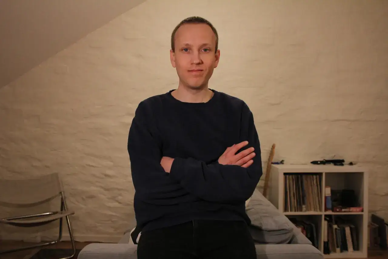 Paolo Nobis Sandén sitter med armarna i kors framför en texturerad vägg. I bakgrunden syns en bokhylla med skivor och böcker.