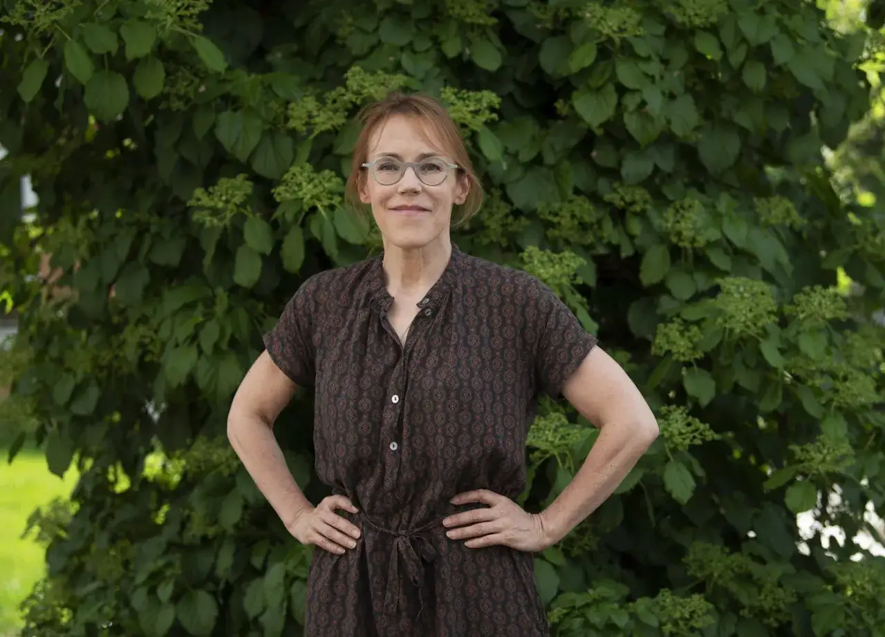 Kvinnan med glasögon, klädd i mönstrad klänning, står framför grönska. Hon påminner om en karaktär från Åsa Larssons familj.