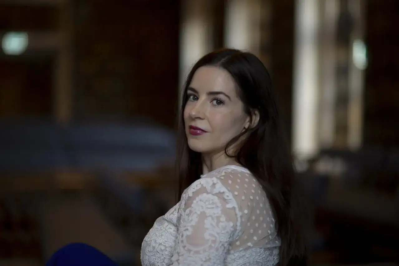 Nina de Geer, med m&ouml;rkt h&aring;r och en vit spetskl&auml;nning, tittar &ouml;ver axeln.