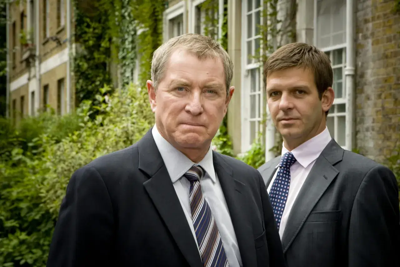Två män i kostym står framför en byggnad med murgröna. De ser ut att vara från grace serie.