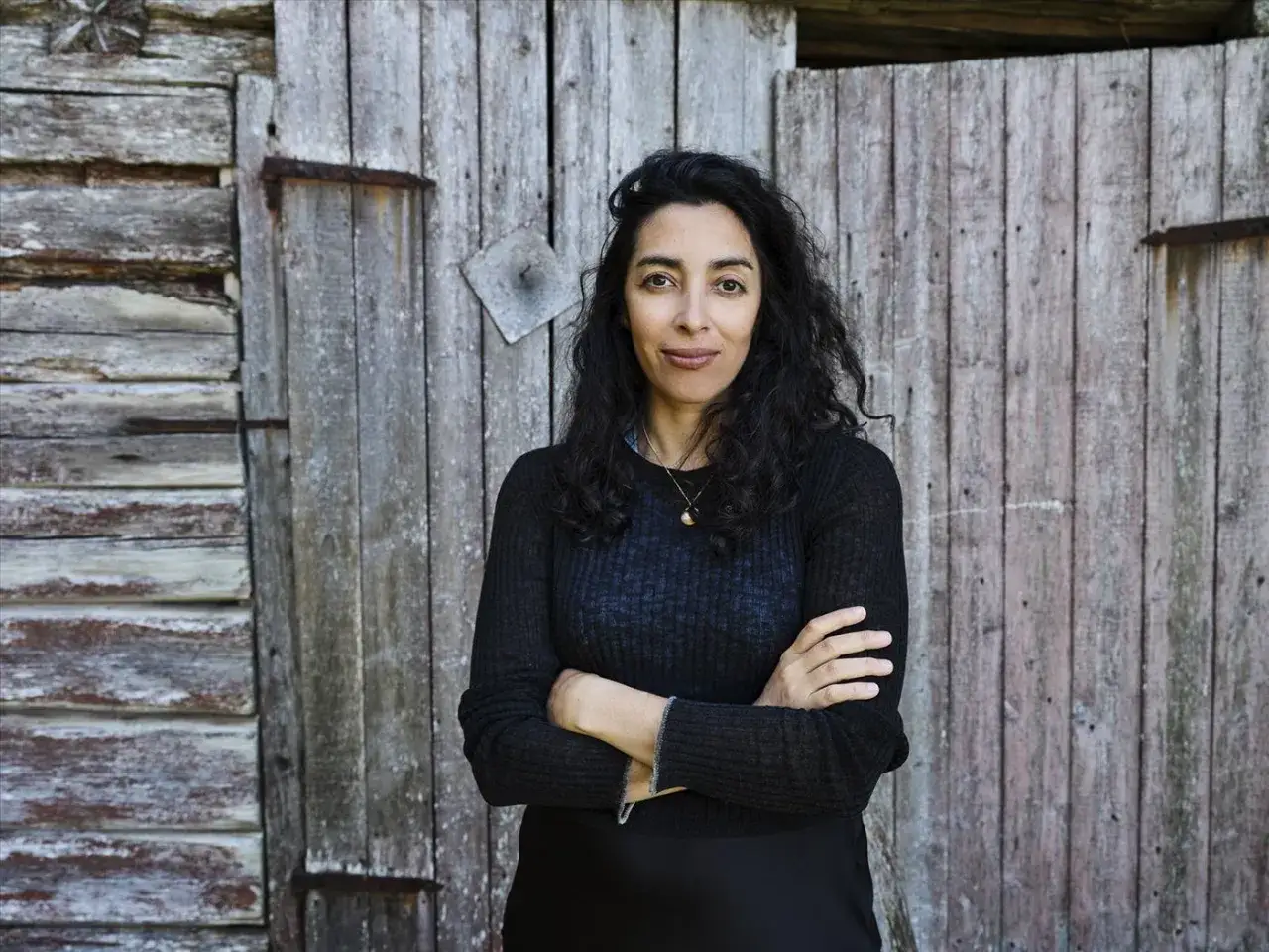 En kvinna med mörkt hår, Negar Naseh, står med armarna i kors framför en sliten trävägg.