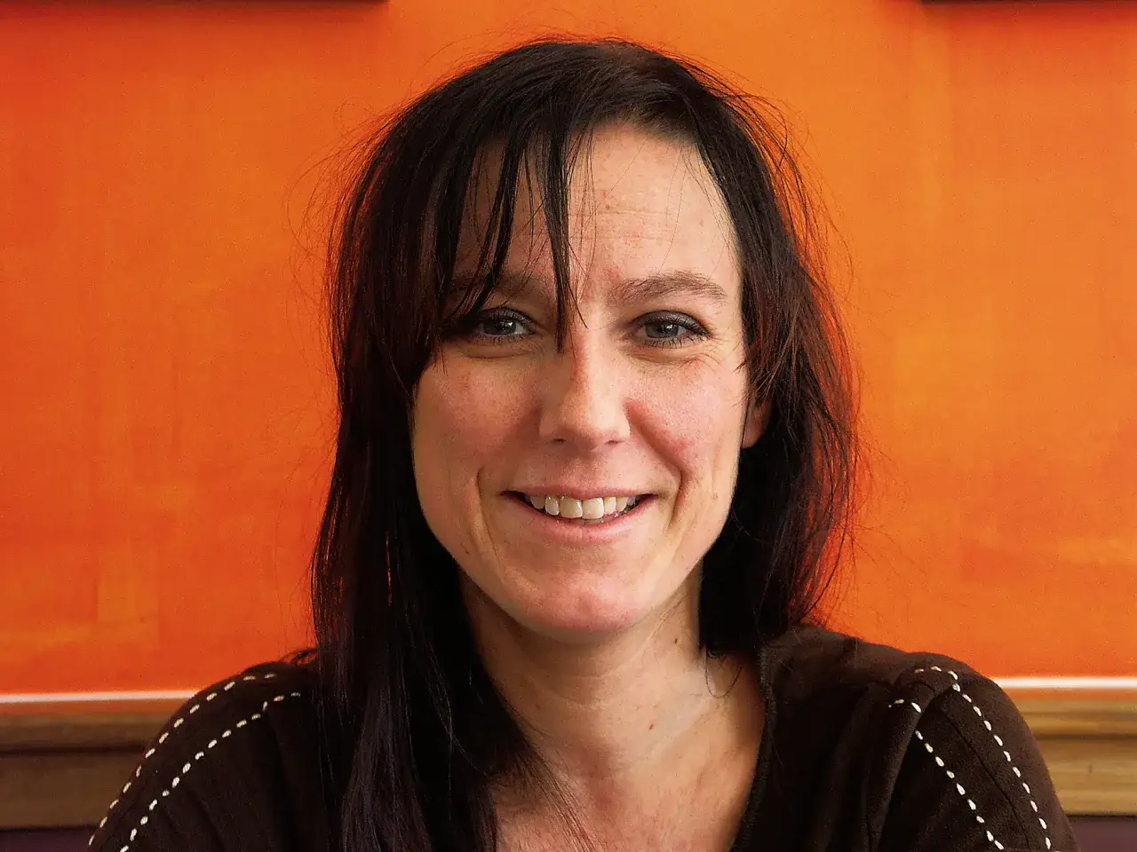 Porträtt av Lena Ollmark med mörkt hår och ett leende, mot en orange bakgrund.