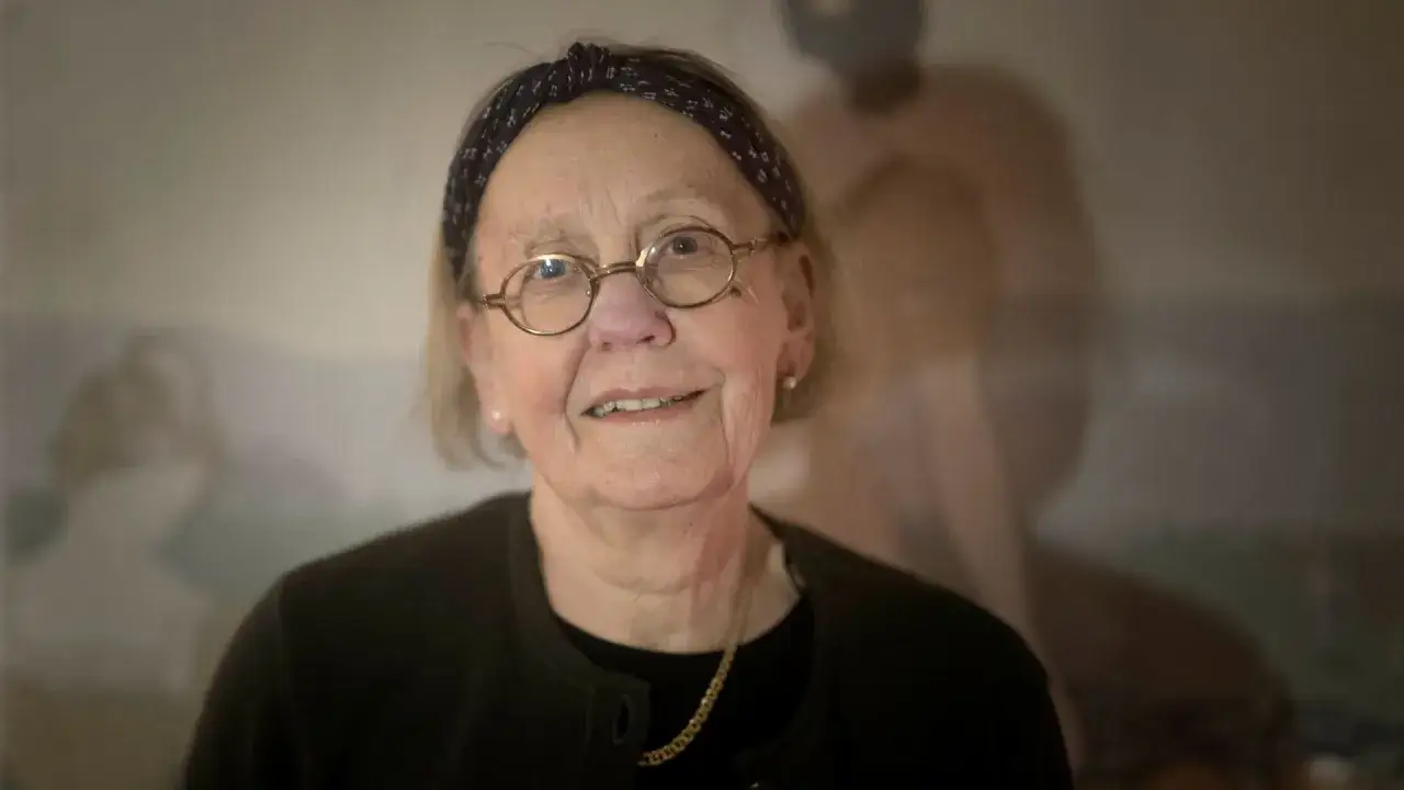 Anna Asp, scenograf, med glas&ouml;gon och pannband, ler mot kameran. Bakgrunden &auml;r suddig med en figur.