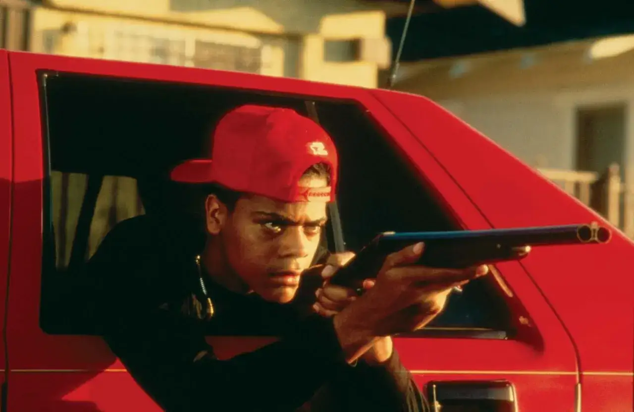 En ung man med röd keps siktar med ett hagelgevär från en röd bil. En scen från rollistan i Boyz n the Hood.