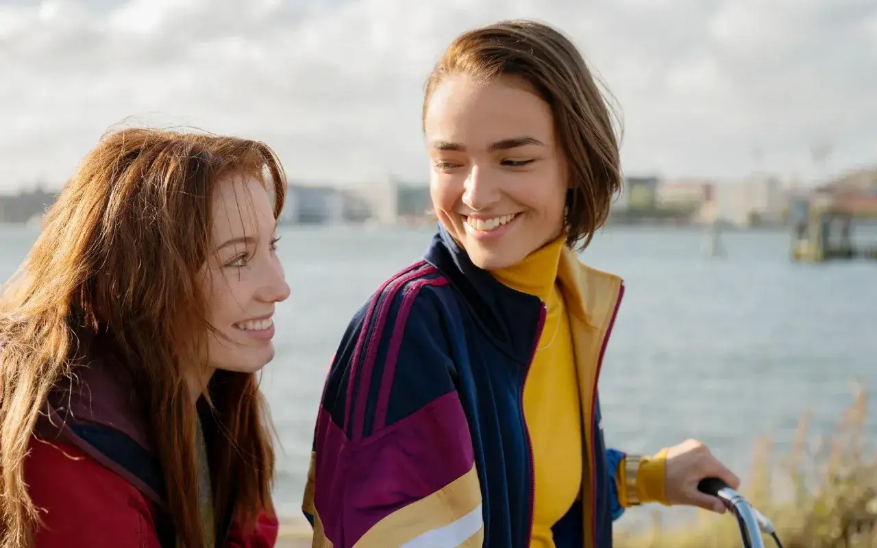 Två unga kvinnor ler mot varandra vid vattnet, en scen som påminner om en svensk gayfilm.