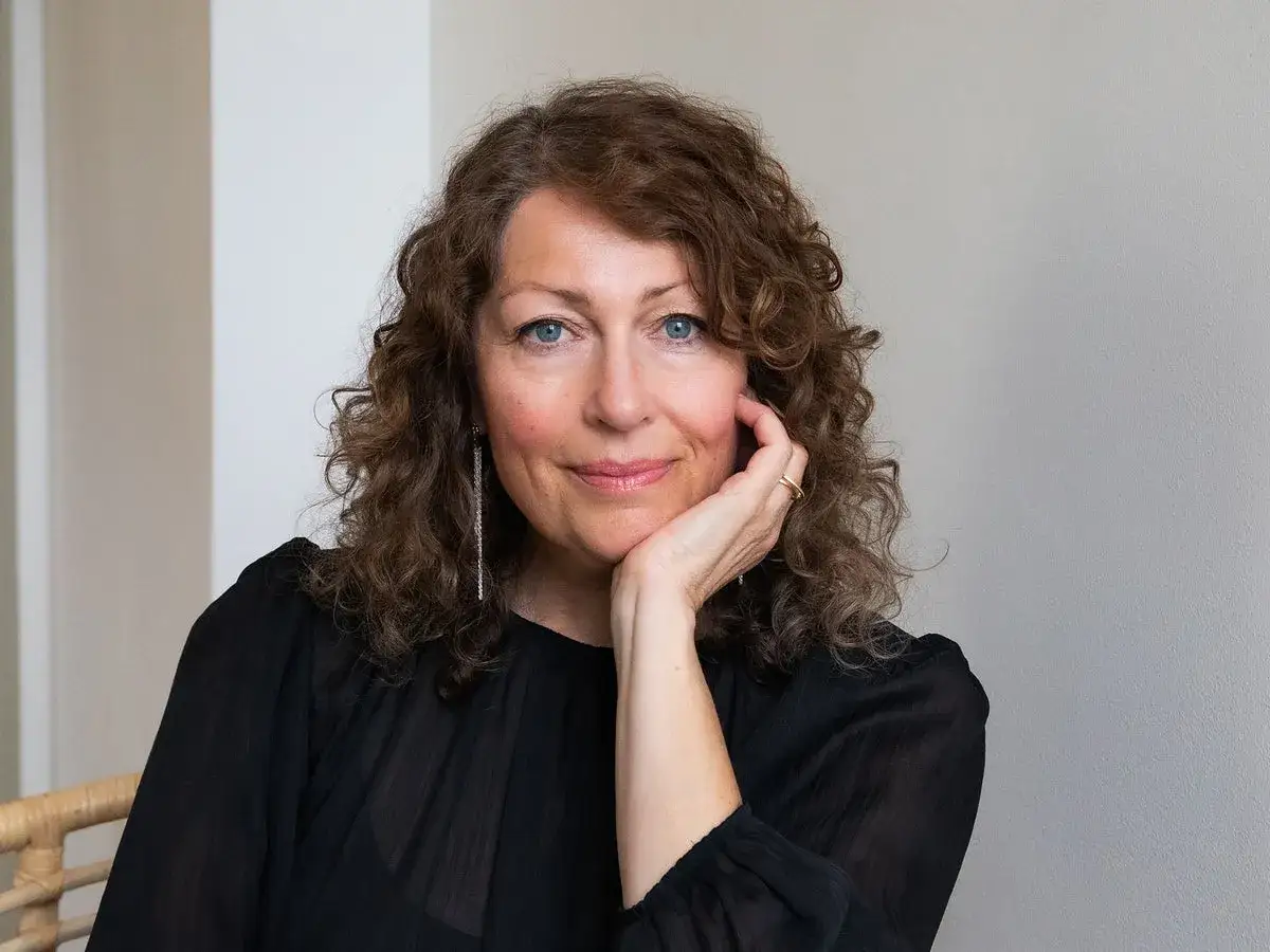 Elisabeth Åsbrink, med sitt lockiga hår och blå ögon, vilar kinden i handen. Hon bär en svart blus och ett smycke.
