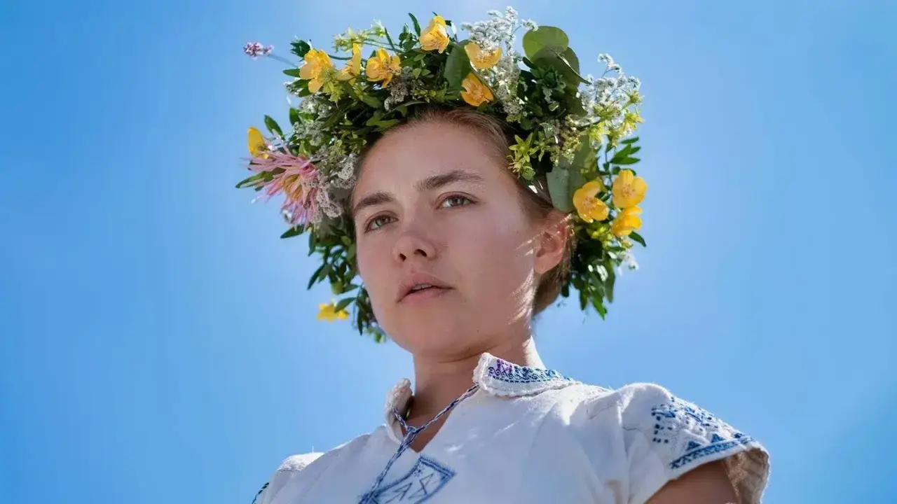 En kvinna med blomsterkrans i håret, klädd i en broderad vit klänning, en scen från filmen Midsommar mot en klarblå himmel.