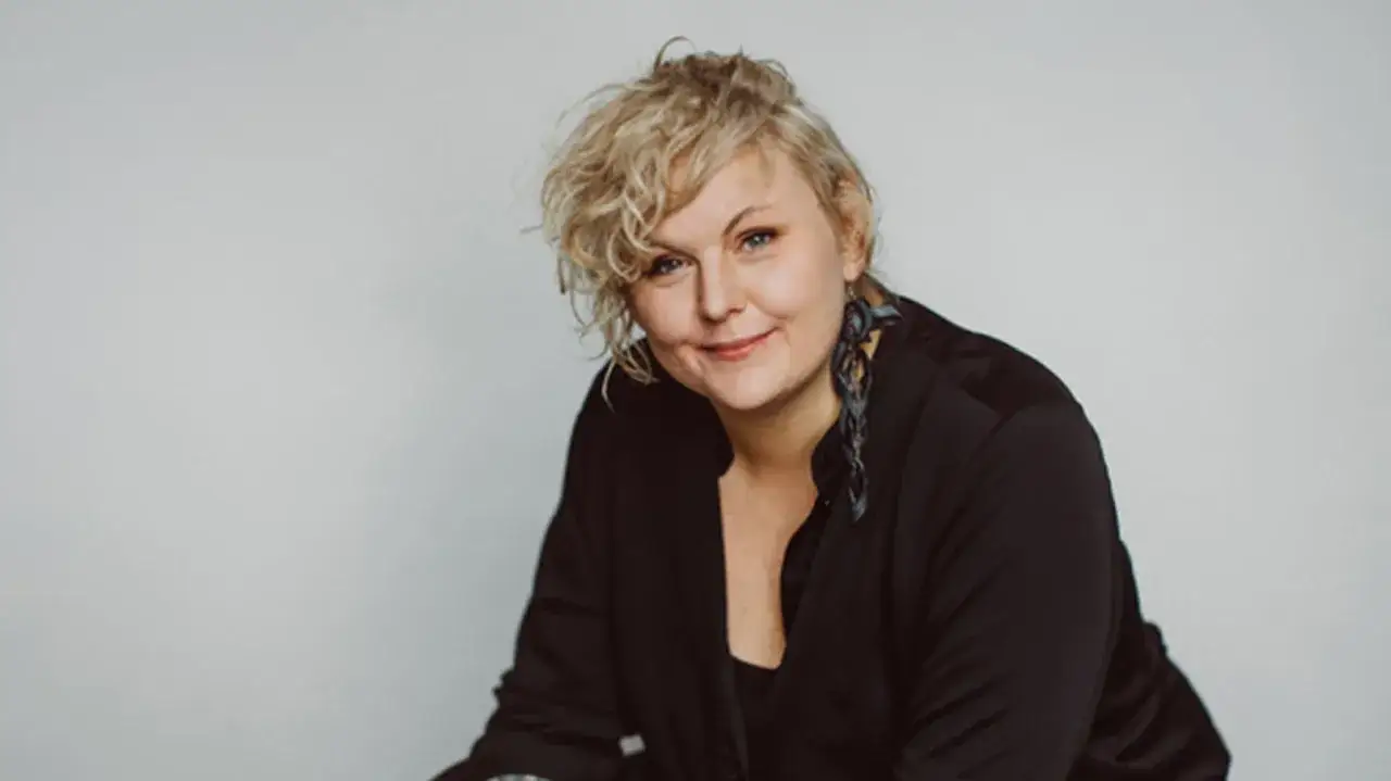 Johanna Thydell, med sitt lockiga blonda h&aring;r och stora &ouml;rh&auml;ngen, ler mot kameran i en svart skjorta.