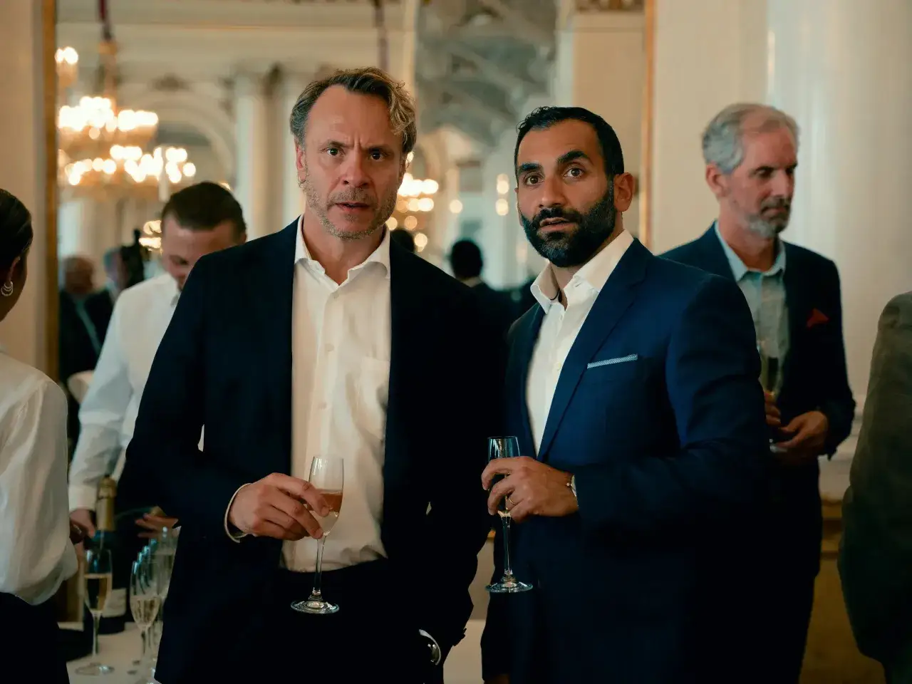Tv&aring; m&auml;n i kostym h&aring;ller glas med champagne. En av dem ser bekymrad ut, som om han precis sett hundarna p&aring; SVT.