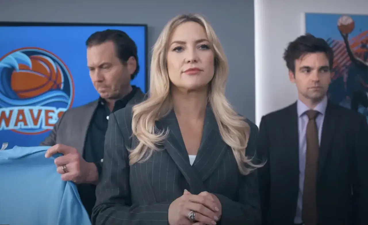 Kate Hudson och två män står framför en skärm med en basketboll och vågor. En man håller upp en ljusblå tröja. De verkar vara på en running point för ett sportlag.